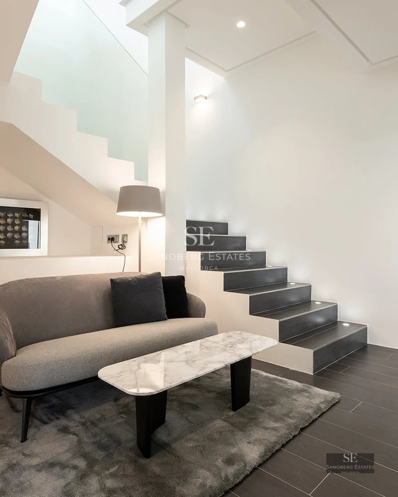 Modern minimalist lounge area with a grey sofa, marble coffee table, and a sleek dark staircase against white walls.