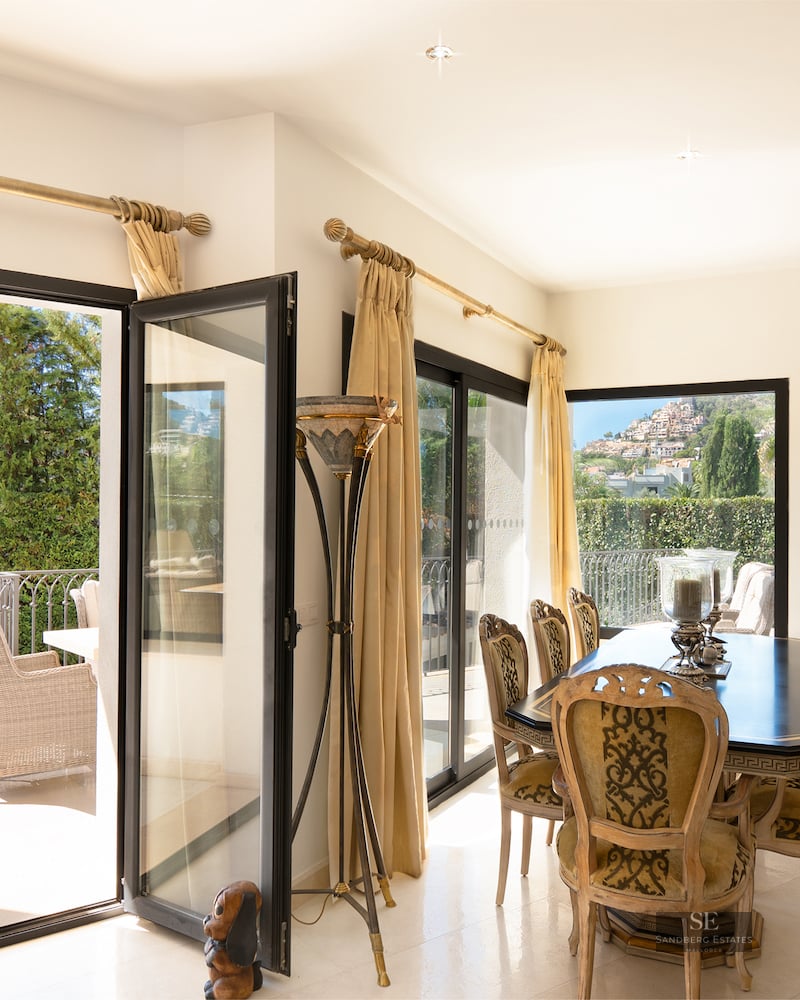 Luxury dining room featuring a black table, ornate chairs, and large open glass doors leading to a balcony with lush greenery.