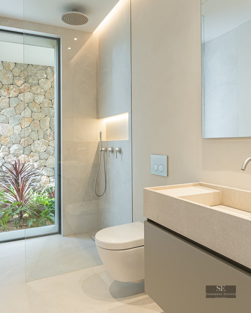 Minimalist bathroom with a stone double vanity, walk-in shower, and floor-to-ceiling window facing a natural stone wall.