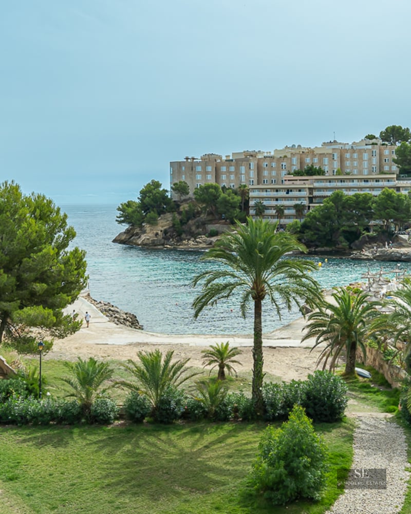 A lush garden with palm trees and a stone path overlooking a turquoise Mediterranean bay and coastal buildings.