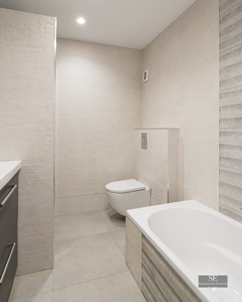Salle de bain moderne avec baignoire blanche, WC suspendu et meuble gris sur fond de carrelage beige texturé.