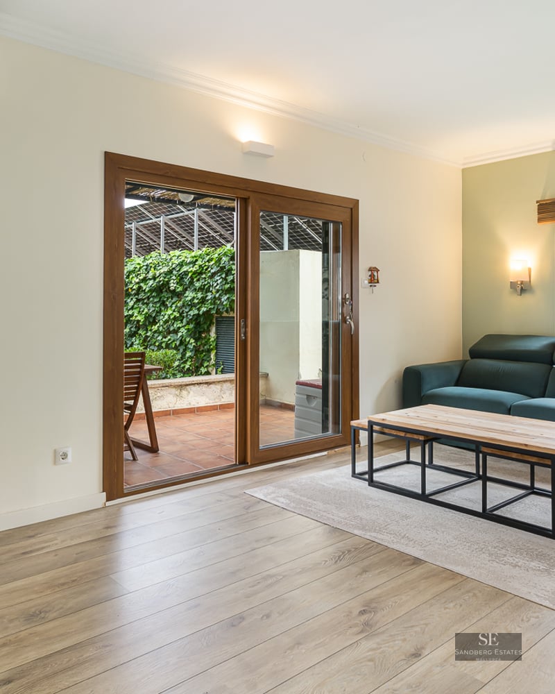 Bright living room with teal sofa, armchair, wood floors, and glass doors leading to a lush green terrace.