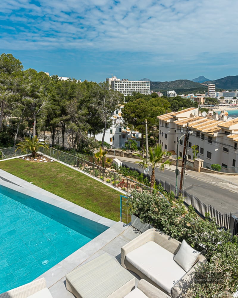 A long turquoise swimming pool on a terrace with glass railings, overlooking a coastal town and distant mountains.