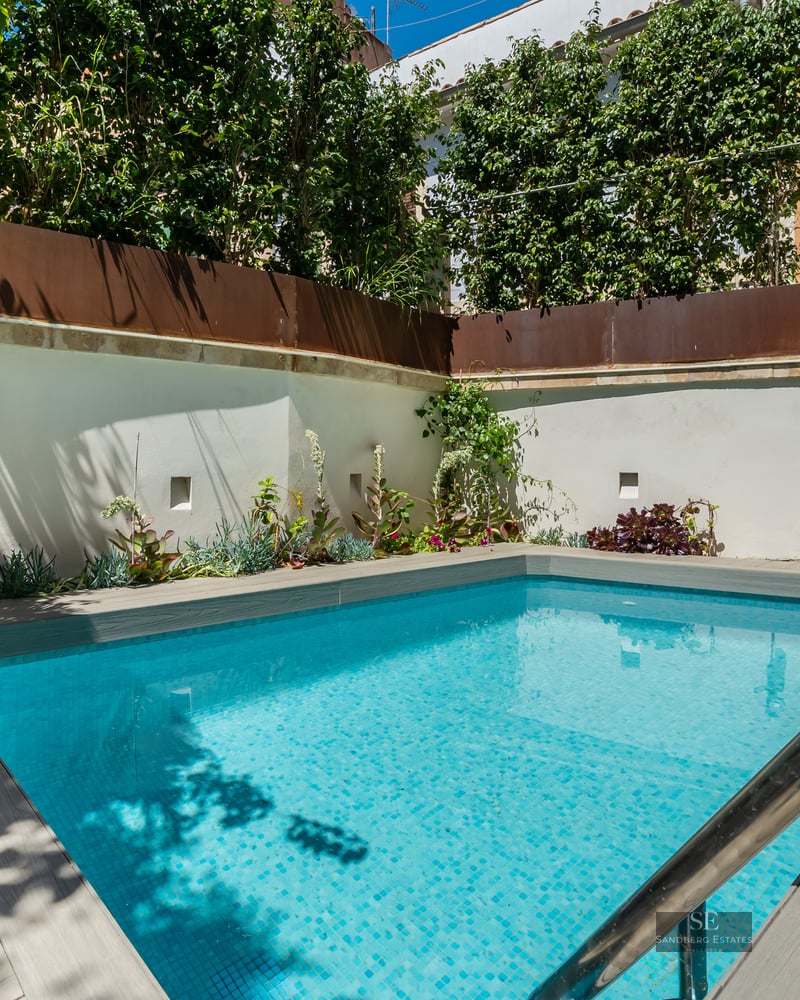 Small turquoise swimming pool with grey wooden decking surrounded by white walls and lush green plants.