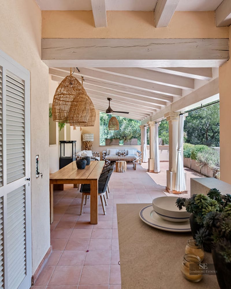 Large covered terrace with terracotta floors, exposed beams, a long wooden dining table, and wicker pendant lights.