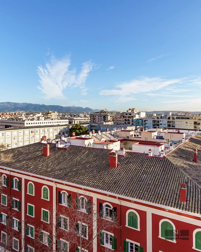High-angle city view featuring a red building with tiled roofs, urban streets, and distant mountains under a clear sky.