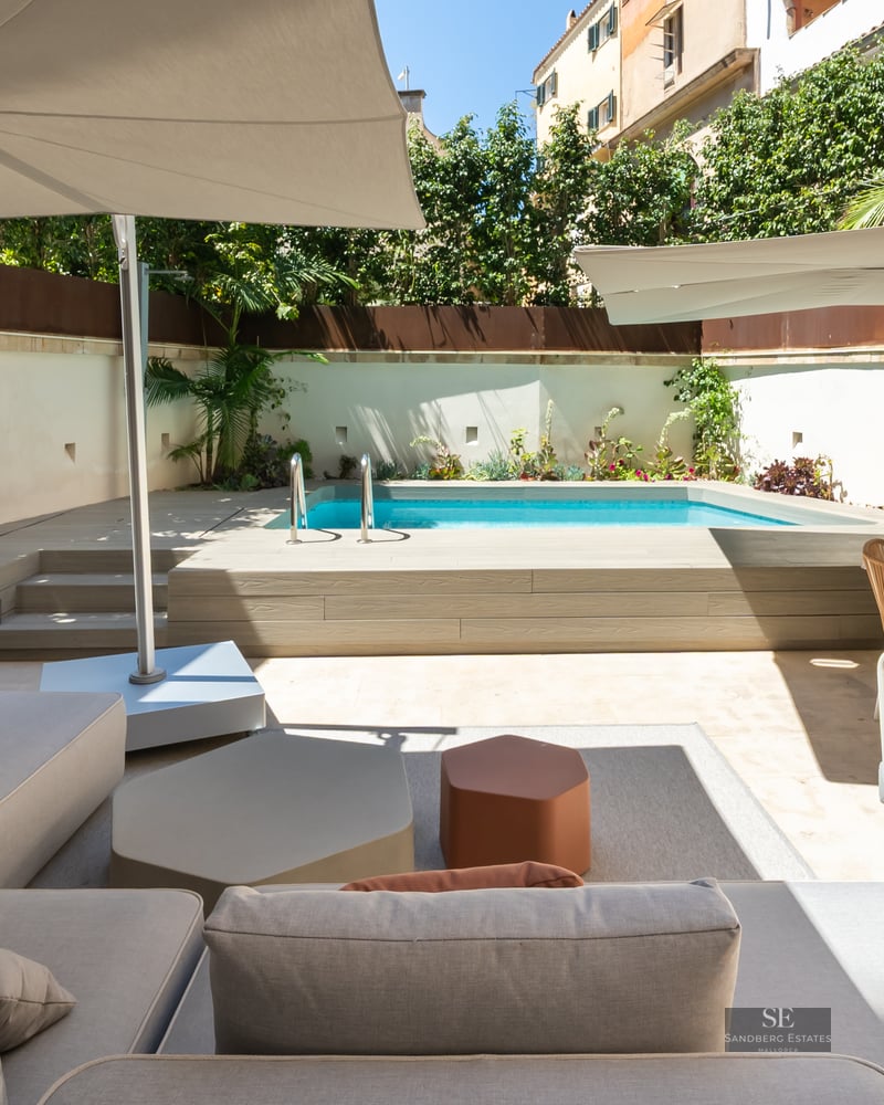 Modern terrace featuring a turquoise pool, wooden deck, grey sofas, and sun umbrellas under a clear sky.