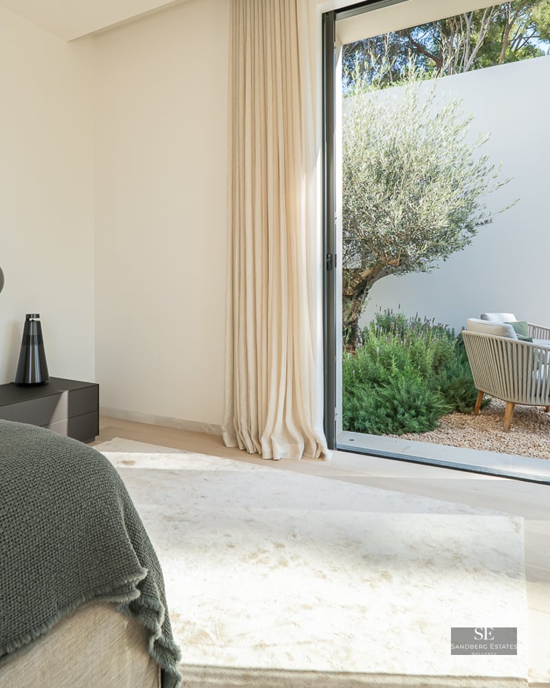 Modern bedroom with a large bed, designer pendant lights, and sliding glass doors opening to a sunlit garden with an olive tree.