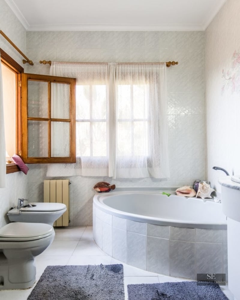 Spacious bathroom featuring a large corner bathtub, marble-topped vanity, toilet, bidet, and wooden-framed windows.