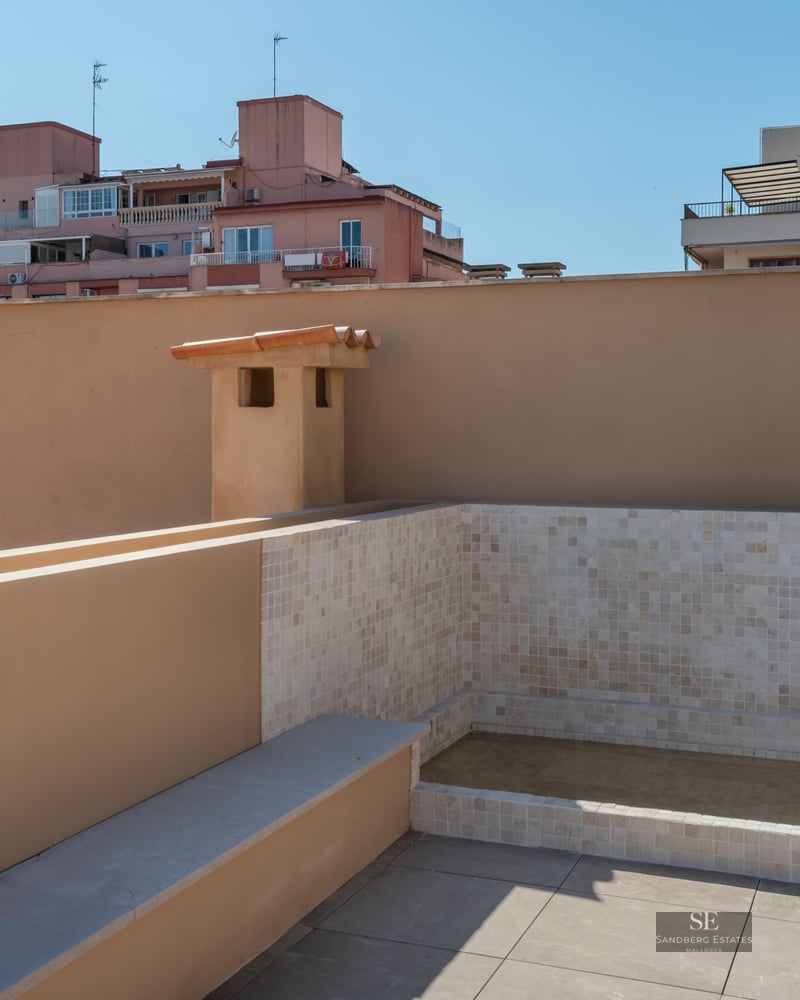A modern rooftop terrace featuring a tiled plunge pool, beige stone flooring, and views of city buildings under a blue sky.