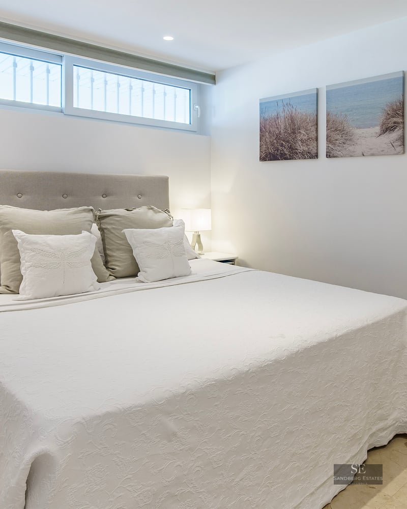 A bright bedroom featuring a large bed with a tufted headboard, white bedding, and coastal artwork on the wall.