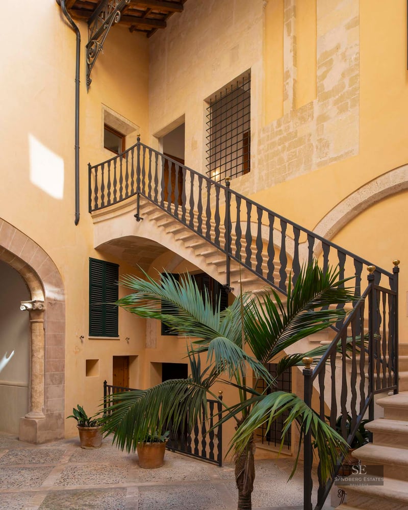Traditional courtyard with yellow walls, a stone staircase with iron railings, and a central palm tree.