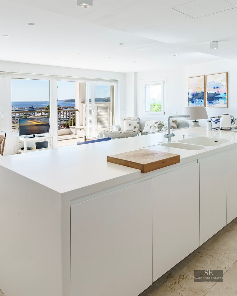 Minimalist white kitchen island leading to a dining area with a panoramic view of the harbor and sea.