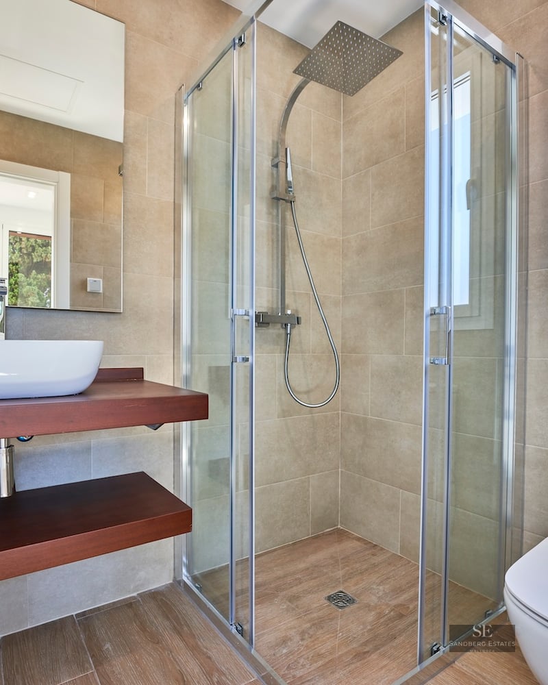 Modern bathroom with beige tiles, glass walk-in shower, wooden floating vanity, vessel sink, and white toilet.
