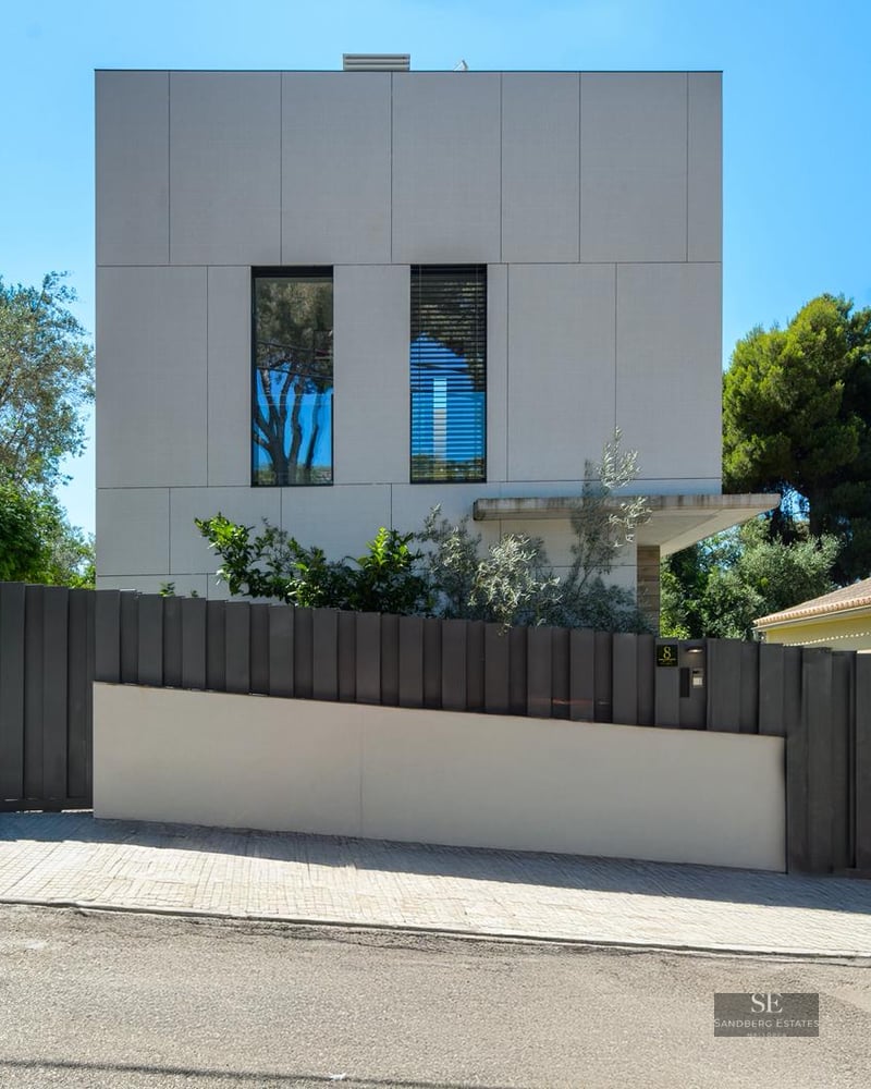 Exterior view of a modern white cubic house with a dark grey metal fence and green trees under a clear blue sky.