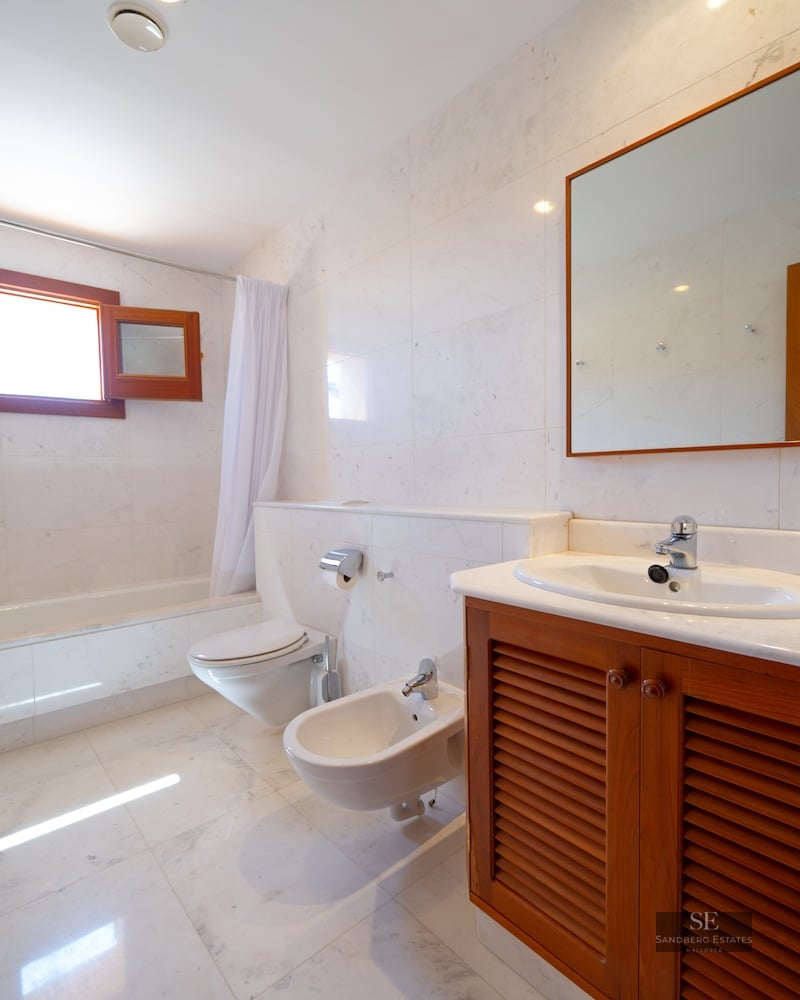 Bathroom featuring white marble walls and floors, a wooden vanity, toilet, bidet, and bathtub under a window.