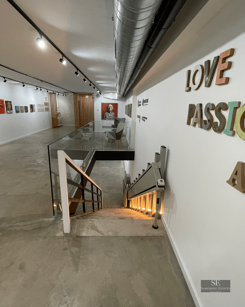 Modern art gallery with colorful paintings, concrete floors, a glass staircase, and a wheelchair lift.