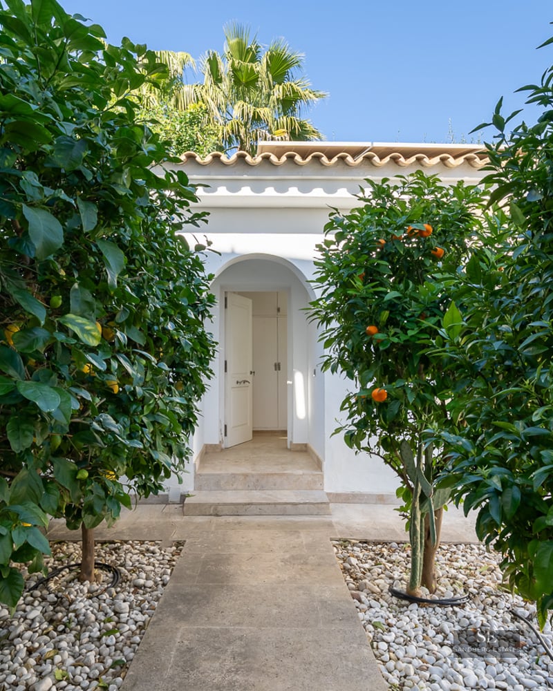 A stone path lined with lemon and orange trees leads to an arched white villa entrance under a clear blue sky.