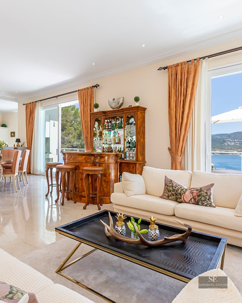 Spacious living room with cream sofas, classic wooden bar, dining area, and large glass doors overlooking the sea.