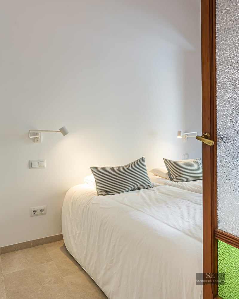 Modern bedroom view through a wooden door with textured glass, featuring a white bed and wall-mounted reading lamps.