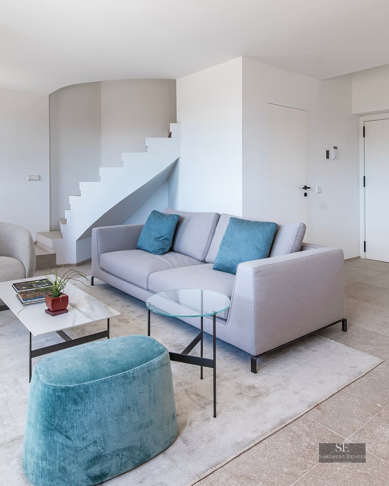 Bright minimalist living room with grey sofa, teal ottoman, white staircase, and marble coffee table.