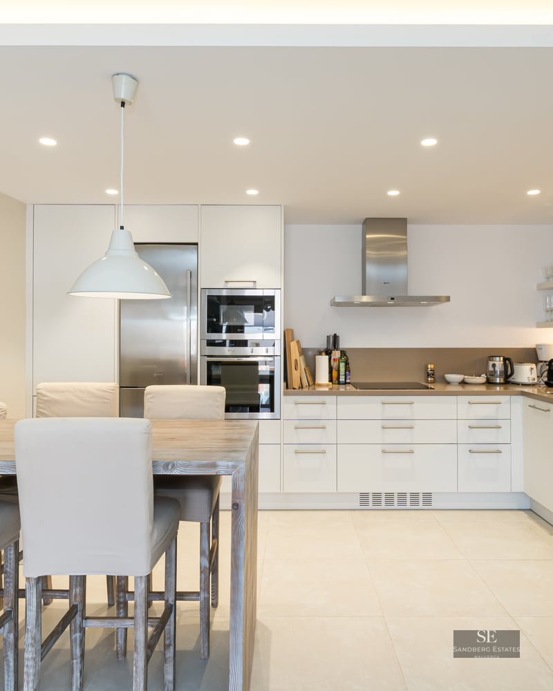 A bright modern kitchen featuring white cabinets, stainless steel appliances, and a large wooden dining table with cream chairs.