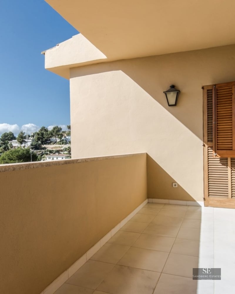 Covered balcony with wooden shutters and tiled floor overlooking the sea and lush greenery.