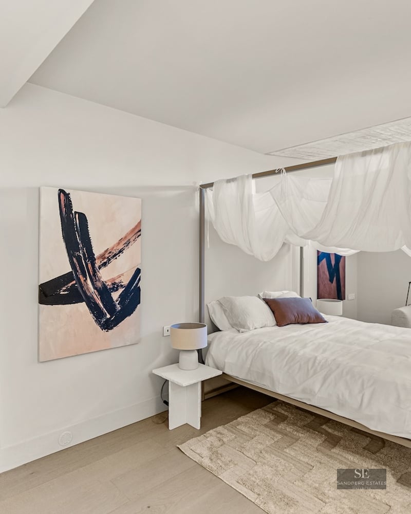 Bright bedroom featuring a four-poster canopy bed with white drapes, abstract art, and light wood flooring.