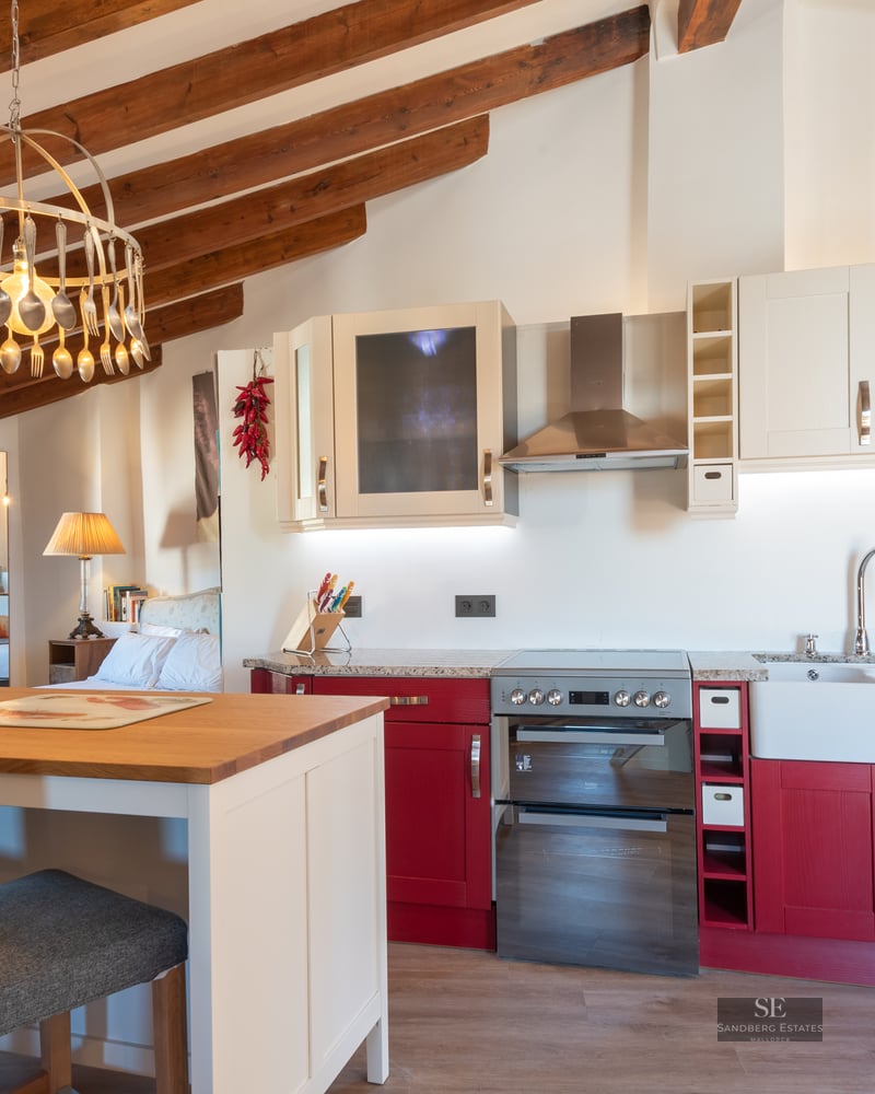 Rustic kitchen featuring red cabinets, stone walls, and exposed wooden ceiling beams with a unique spoon chandelier.