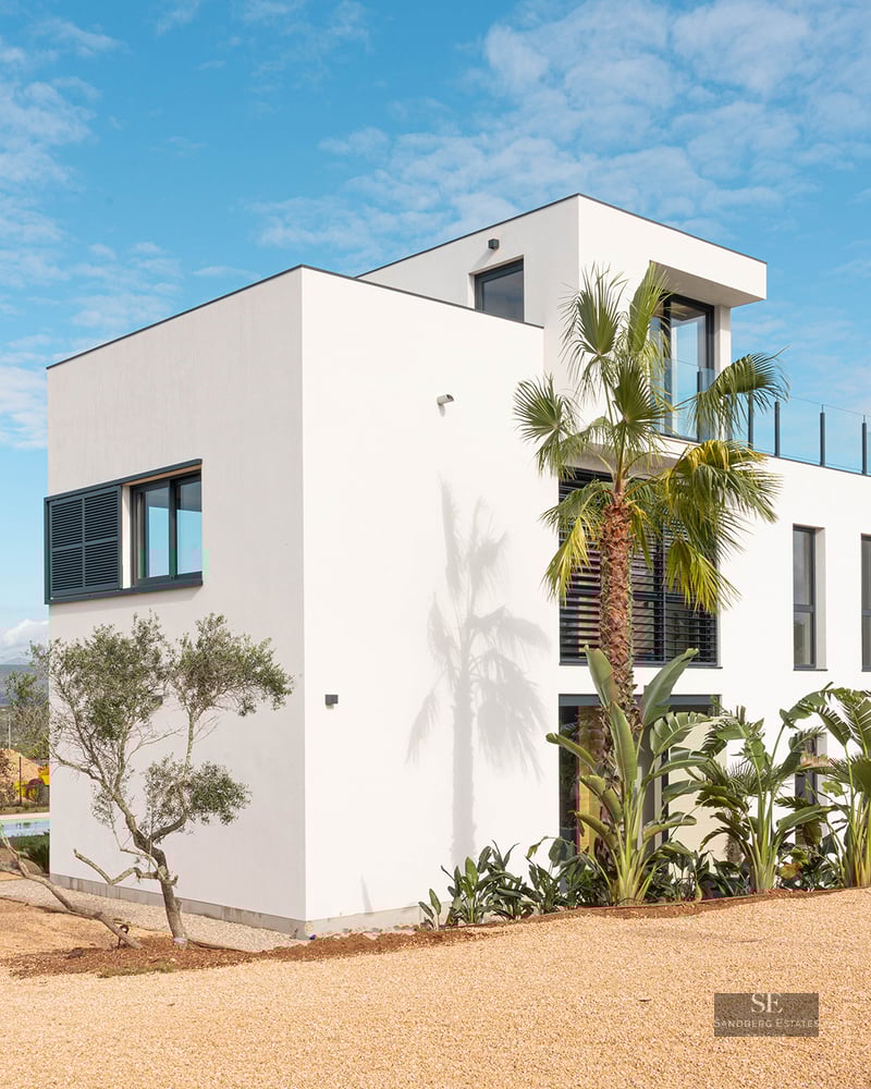 Exterior of a three-level white villa with modern architecture, palm trees, and a gravel yard under a blue sky.