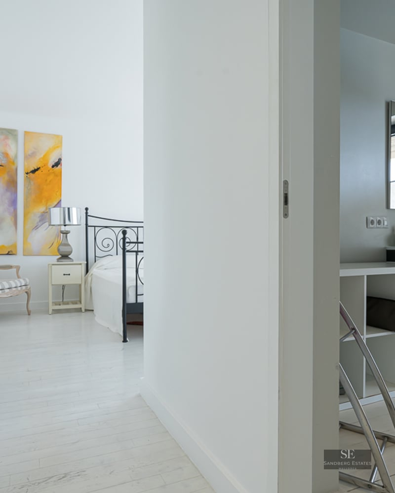 View from a bright hallway into a white bedroom with abstract art and an adjacent bathroom with a black vanity.