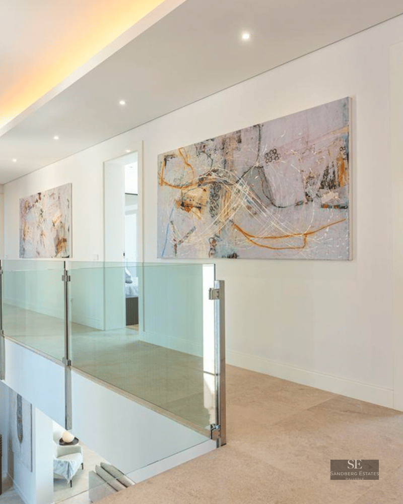 Modern hallway with glass balustrades, abstract art on white walls, and stone floors overlooking a lower level.