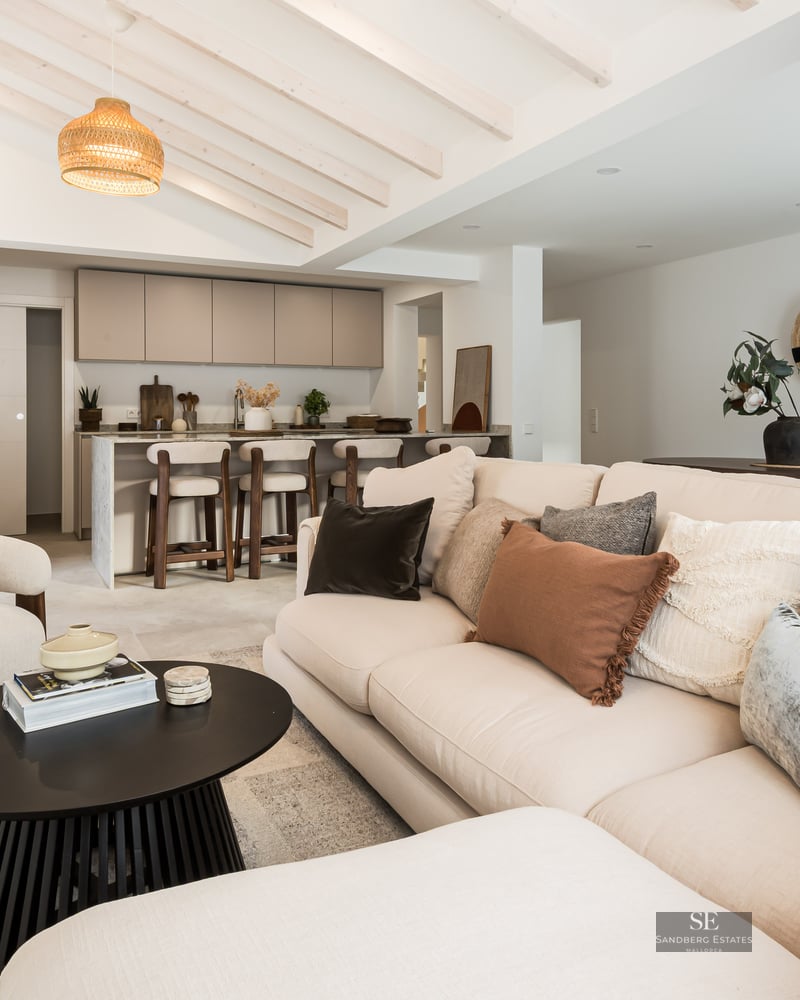 Bright living room with beige sofa, wooden ceiling beams, and a modern kitchen island in the background.