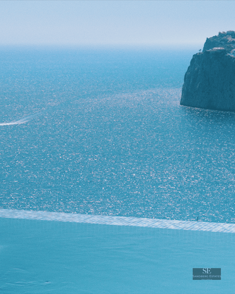 Infinity pool edge overlooking a vast blue sea with a dramatic cliffside coastline and a small boat trail.