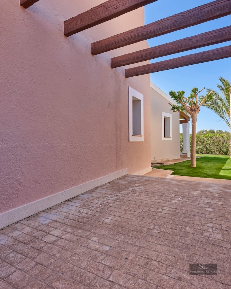 A stone-paved outdoor passage with pink stucco walls and dark wooden beams leading to a green garden.