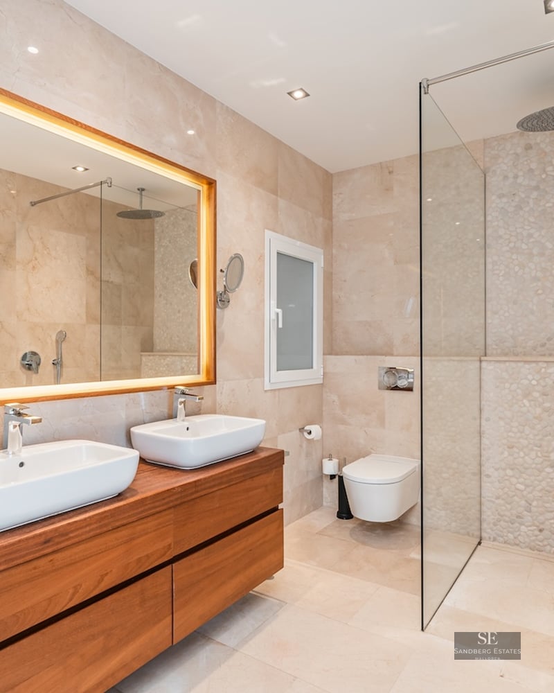 Modern master bathroom with double sinks on wood vanity, backlit mirror, and glass walk-in shower with pebble wall.