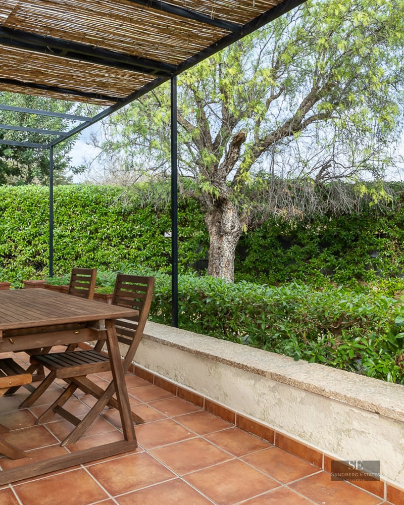 Wooden dining set under a reed pergola on a terracotta tiled terrace surrounded by lush greenery and a large cactus.
