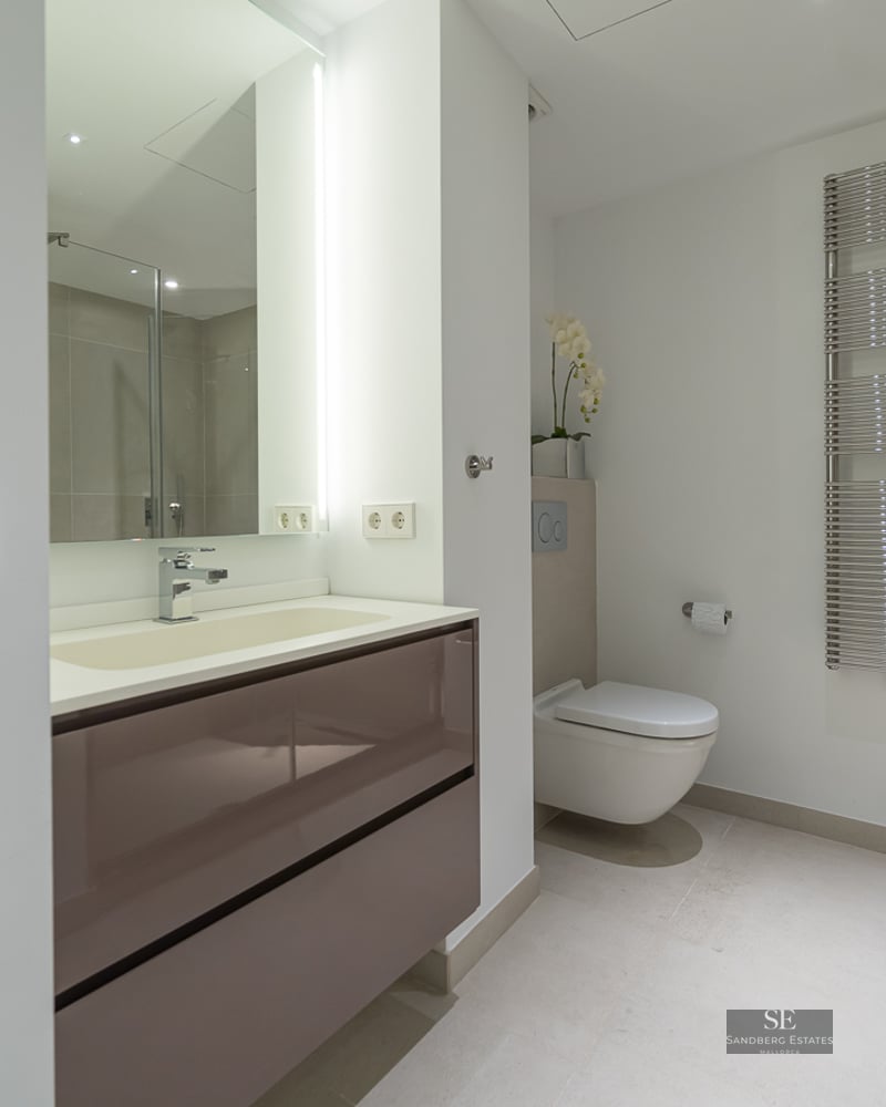 Contemporary bathroom featuring a taupe vanity, illuminated mirror, wall-hung toilet, and chrome heated towel rail.