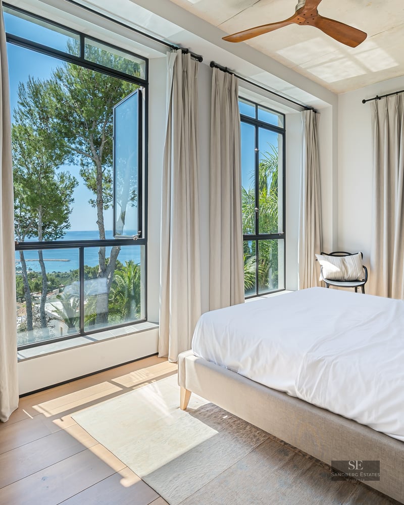 Bright bedroom featuring a white bed, wood floors, a wooden ceiling fan, and large windows with views of the sea and trees.