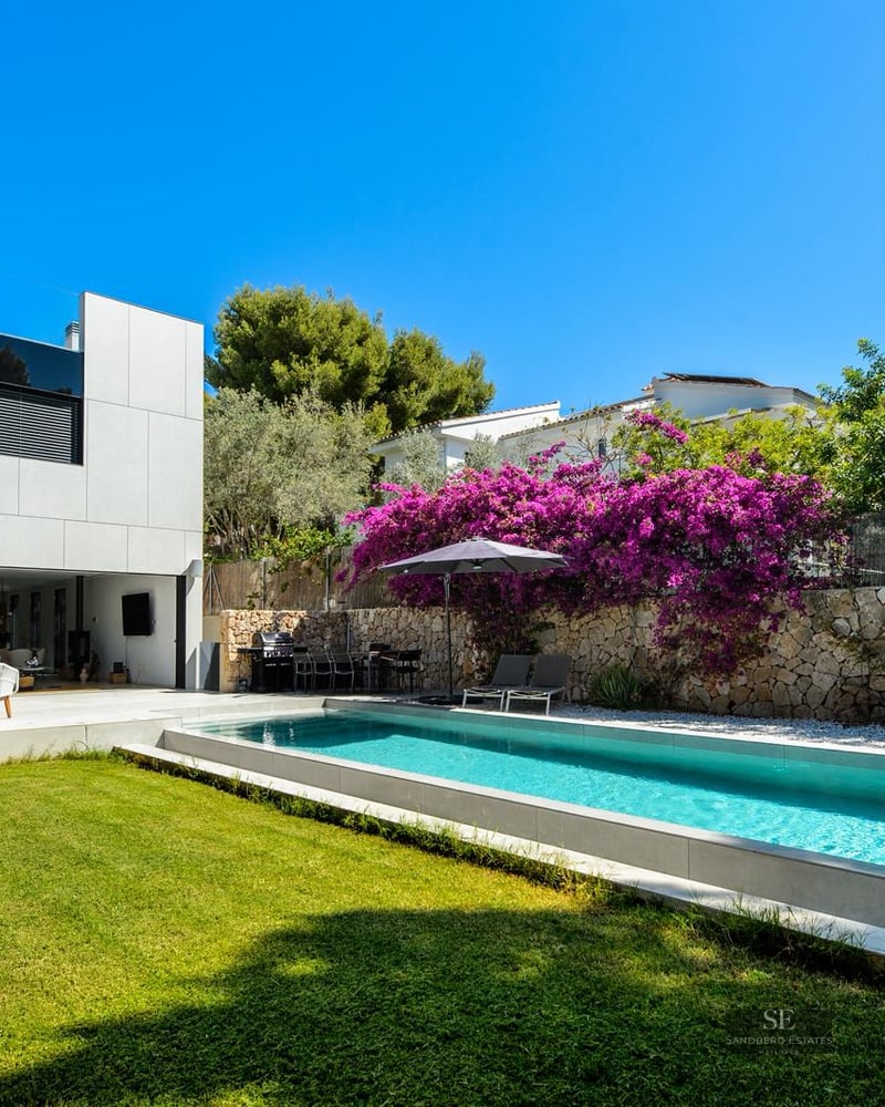 Modern rectangular swimming pool next to a white villa with purple bougainvillea and green lawn under a clear blue sky.
