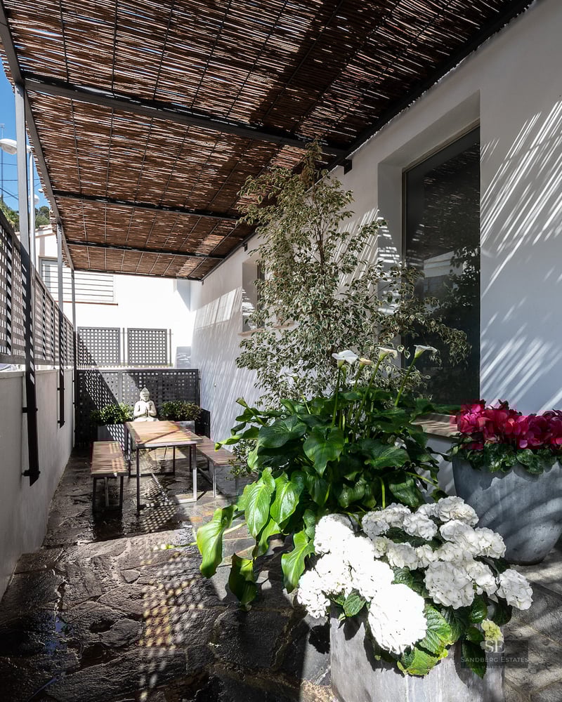 Sunlit terrace with bamboo roof, stone floor, wooden dining set, and lush potted flowers against a white wall.