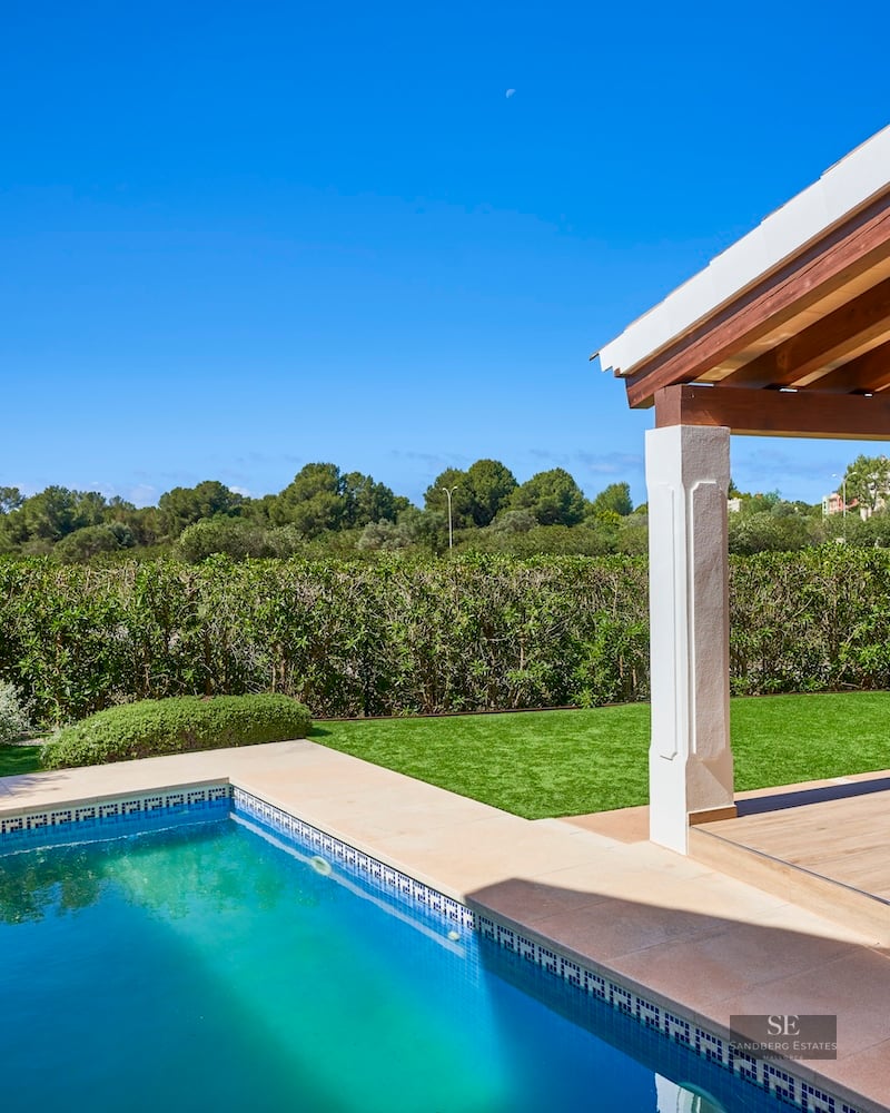 A turquoise swimming pool next to a green lawn and a covered terrace with wooden beams under a clear blue sky.