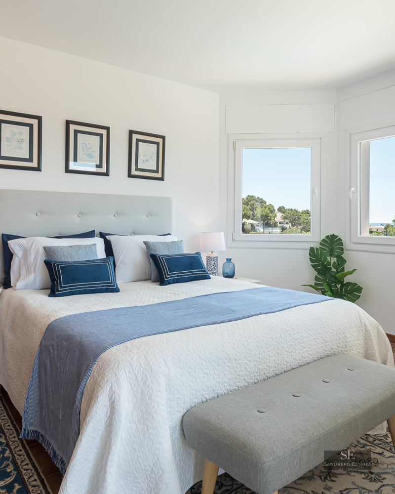 Bright bedroom with a white bed, blue pillows, white dresser, large windows, and terracotta tile floors.