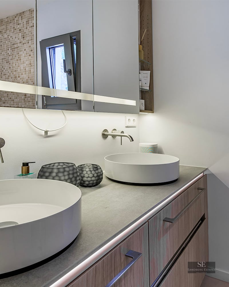 Modern bathroom with two white round vessel sinks on a grey countertop, wood cabinets, and large lit mirror.
