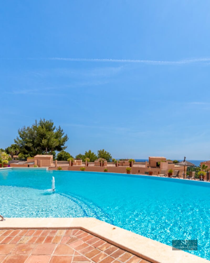 Grande piscine bleue incurvée avec fontaine, terrasse en terre cuite, parasols en paille et vue sur mer.