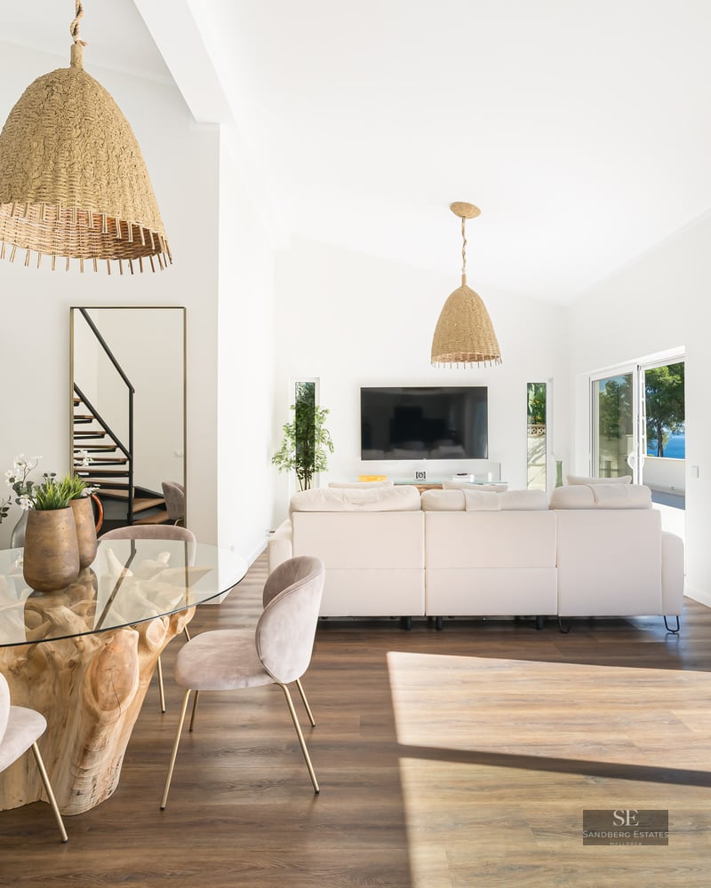 Salon lumineux avec table à manger en bois flotté, canapé blanc et suspensions en osier près de grandes baies vitrées.