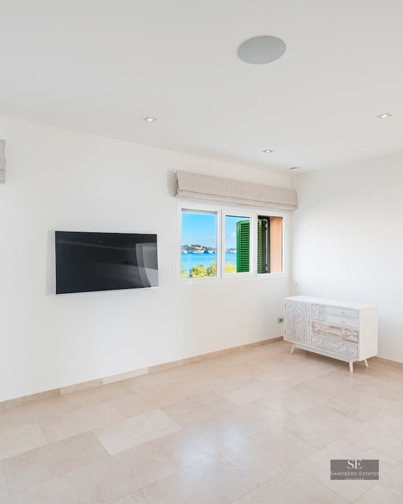 Bright room with white walls, stone floors, wall-mounted TV, and windows overlooking the Mediterranean sea.