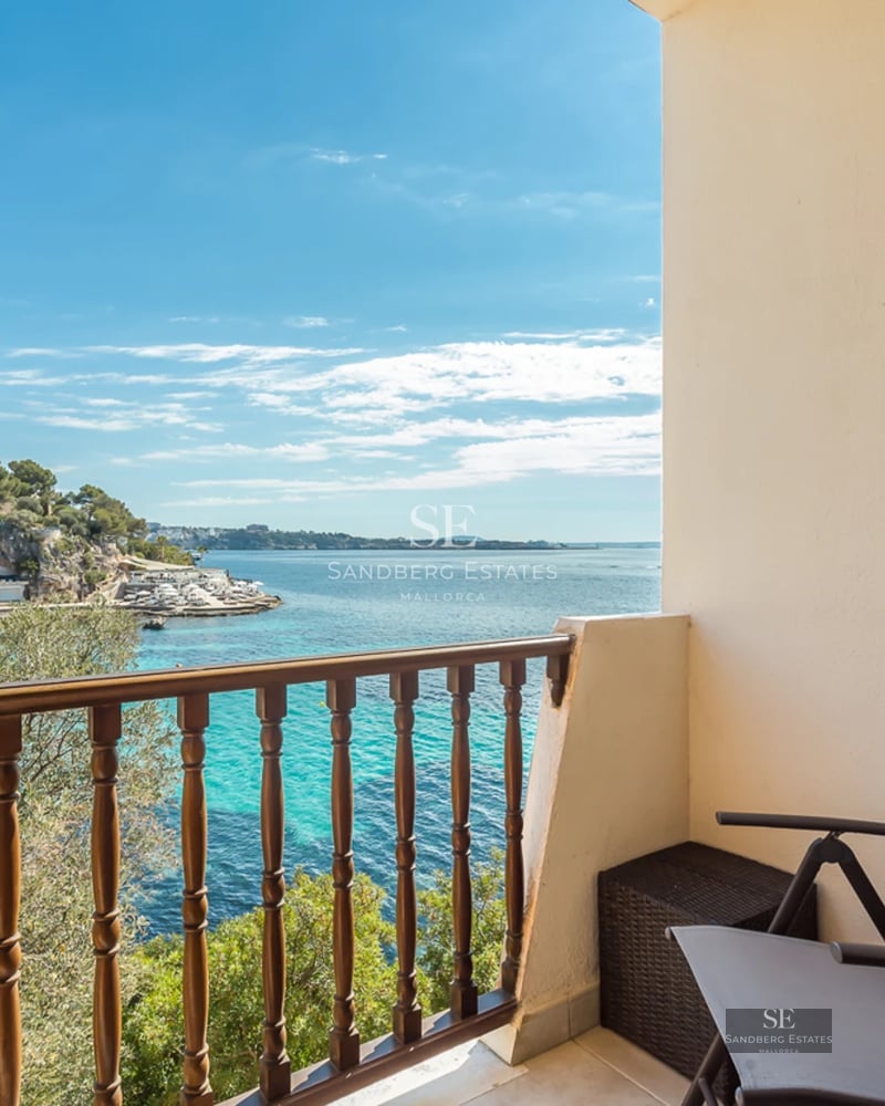 View from a balcony with a wooden railing and grey chair looking over turquoise Mediterranean waters and white buildings.