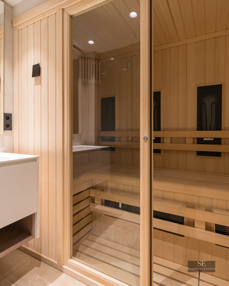 Light wood sauna with glass doors next to a sleek white bathroom vanity and mirror.