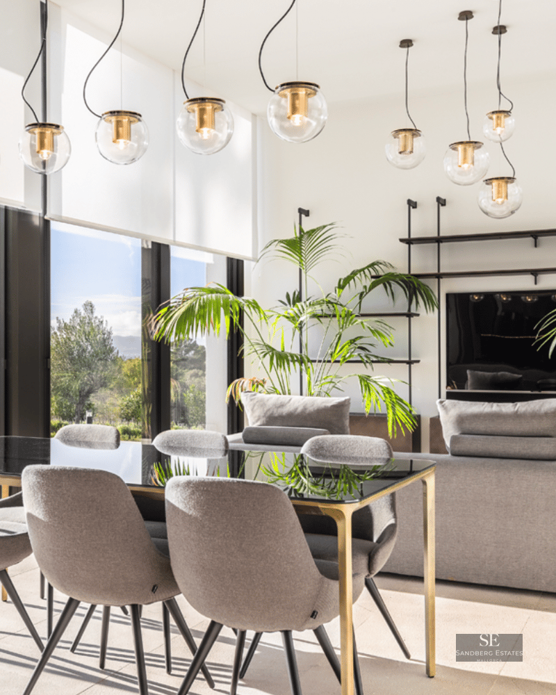 A luxurious modern dining area with a glass table and grey chairs, overlooking a living room and outdoor greenery.